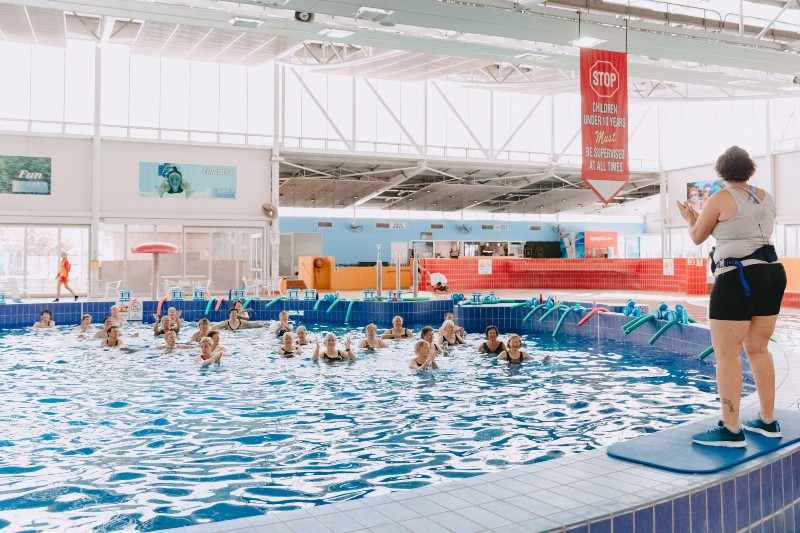A swim instructor guides a diverse group in a pool for an aqua aerobics class. Participants smile and clap, creating an energetic and joyful atmosphere.