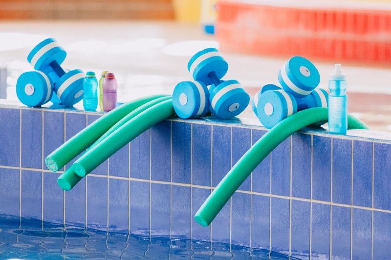 Poolside scene with blue foam dumbbells, green pool noodles, and small plastic bottles on a tiled edge. Bright, inviting, and ready for water exercise.